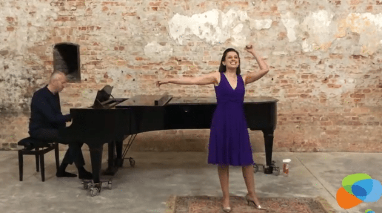 Megan Soprano sings in front of a grand piano with her arms thrown in the air.