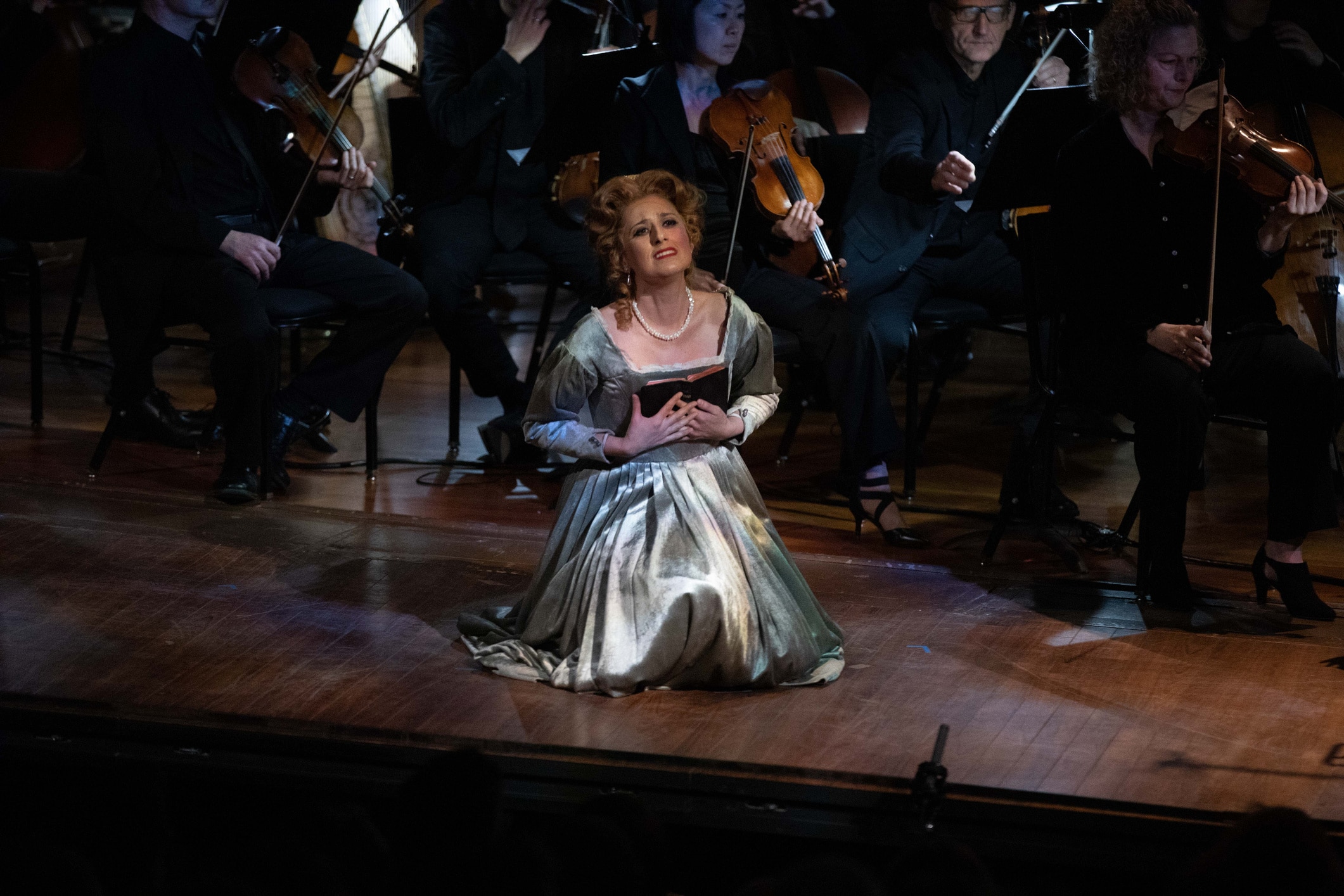Soprano Megan Pachecano kneels at the front of the stage at New England Conservatory's Jordan Hall in Boston, Massachusetts before the Boston Modern Orchestra Project and next to Conductor Gil Rose. They are performing Arnold Rosner's opera The Chronicle of Nine: The Tragedy of Jane Grey. Pachecano wears a shimmering green dress as the 15-year old Lady Jane Grey and clutches her black bible with a concerned look on her face, singing an aria "Into Thy Hands" which quotes a passage from Luke.