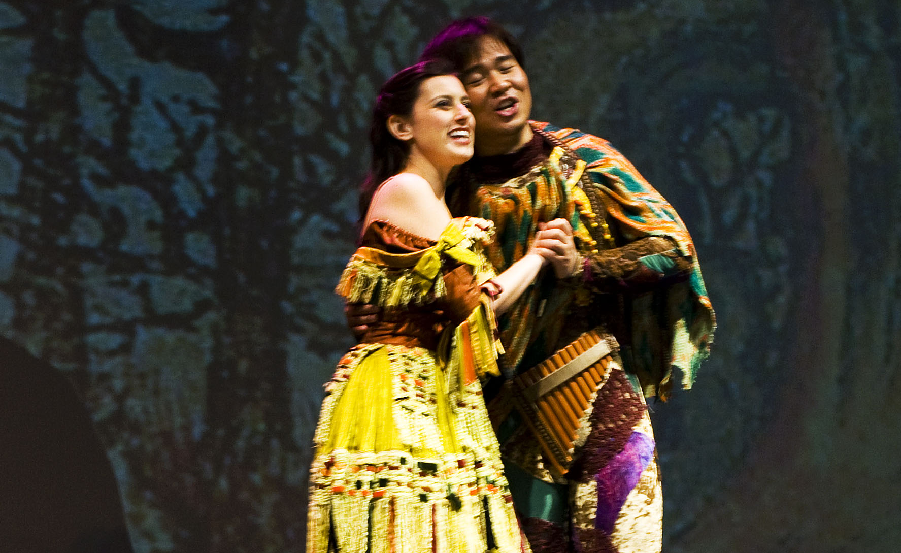 Megan Pachecano and Suchan Kim embrace in a scene from Mozart's Die Zauberflöte with Opera in Williamsburg; they wear colorful costumes reminiscent of bird plumage on stage in front of a set background of trees